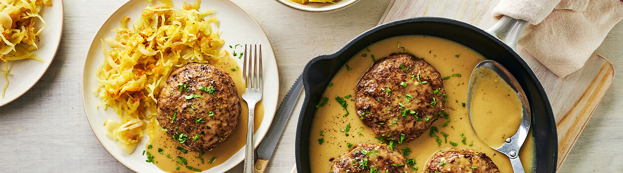 Beef patties in a skillet beside a white plate holding a single beef pattie and cabbage and potato hash